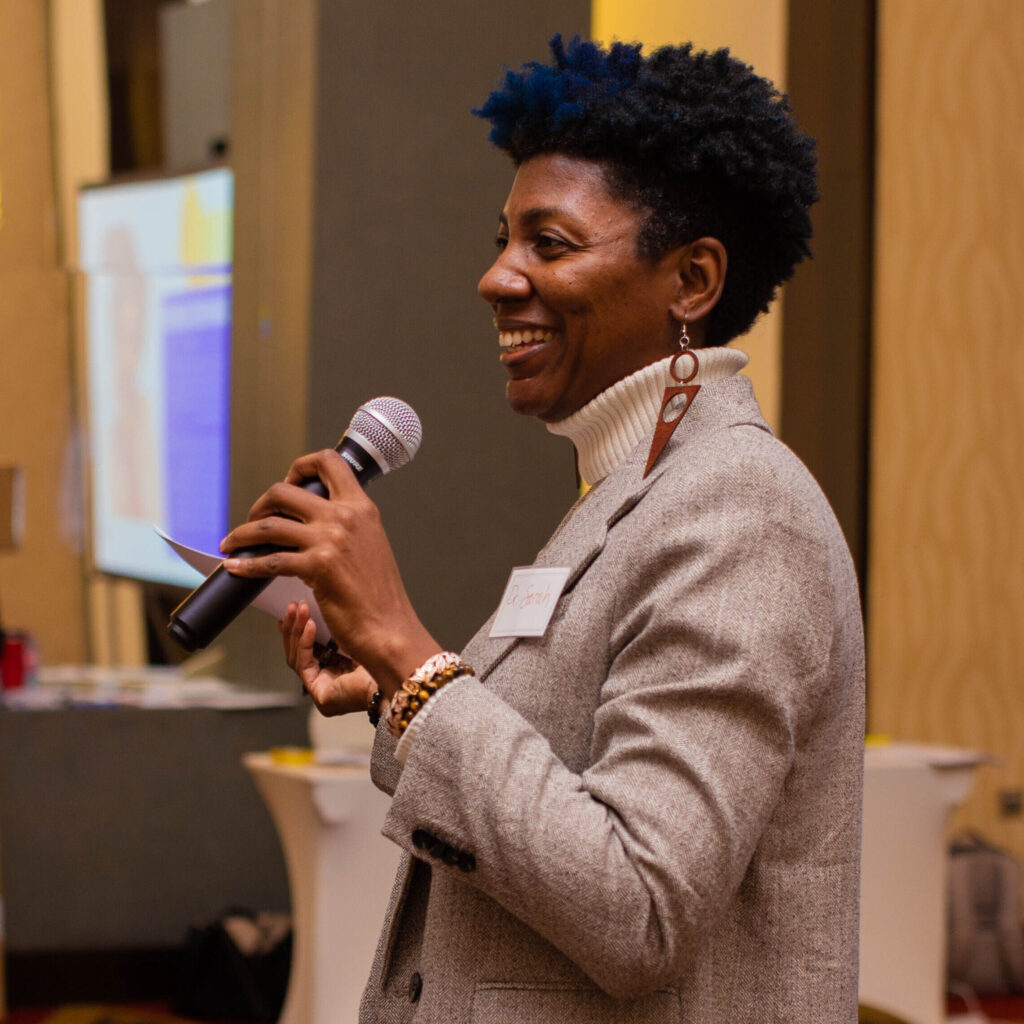 sarah webb smiling and holding a microphone wearing a tan blazer as she delivers a corporate training at a conference hotel in accra ghana