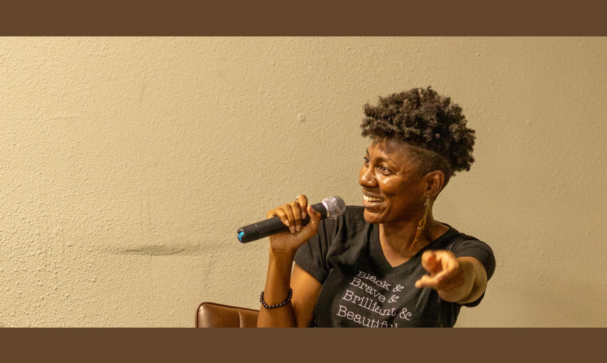 doctor sarah l webb public speaker holding mic while seated on a stool in front of an audience and smiling at her 10 year anniversary event for colorism healing in dallas fort worth texas she's wearing her black and brave and brilliant and beautiful t shirt.